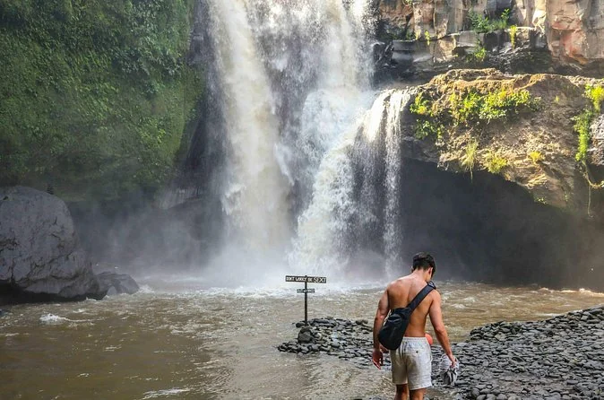 A man wading in the pool near Tegenungan Waterfall with spray behind him